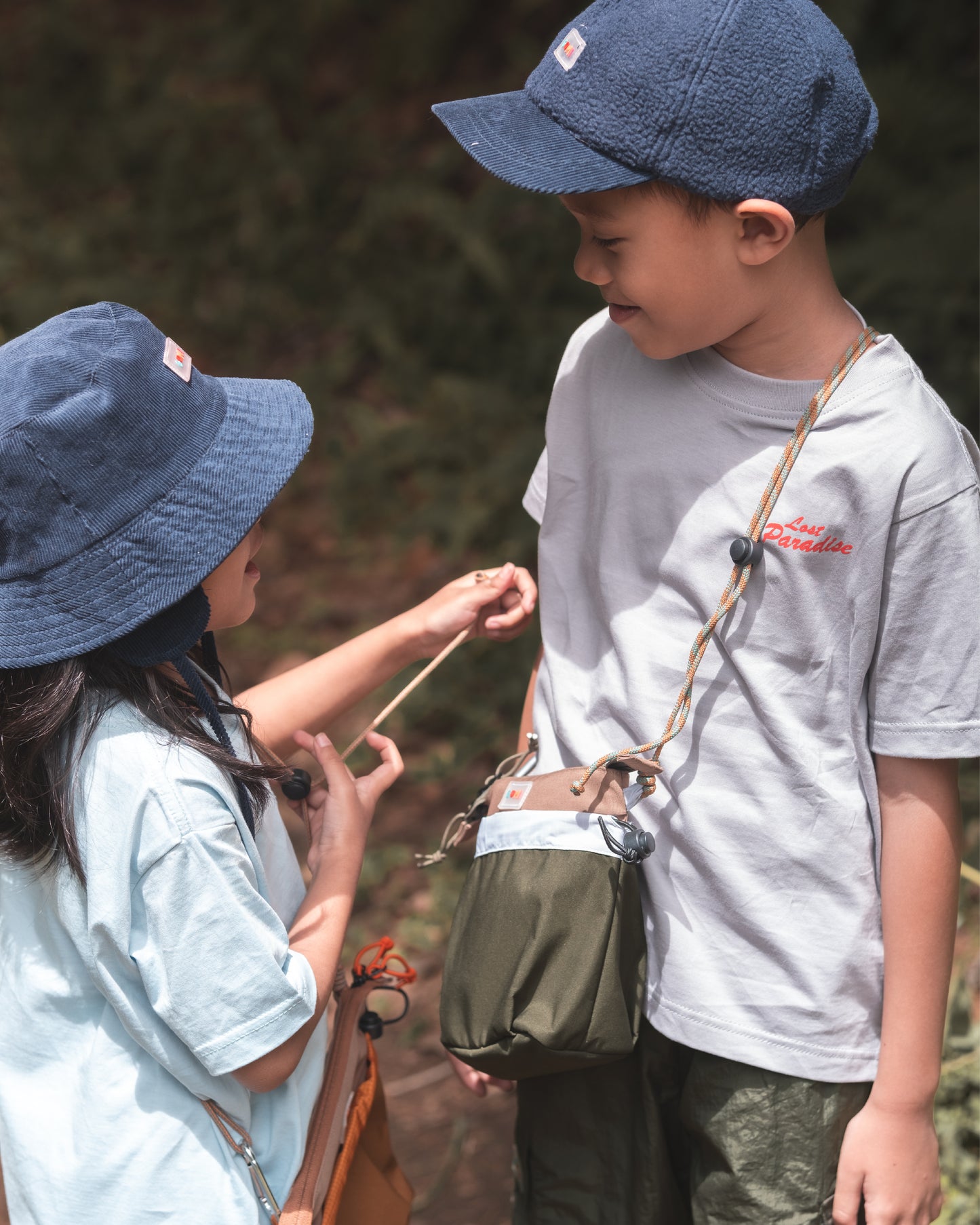 Children wearing lightweight hats and playing outdoors with the PIXIES Nutty Sprout bag, showcasing its adventure-ready design.