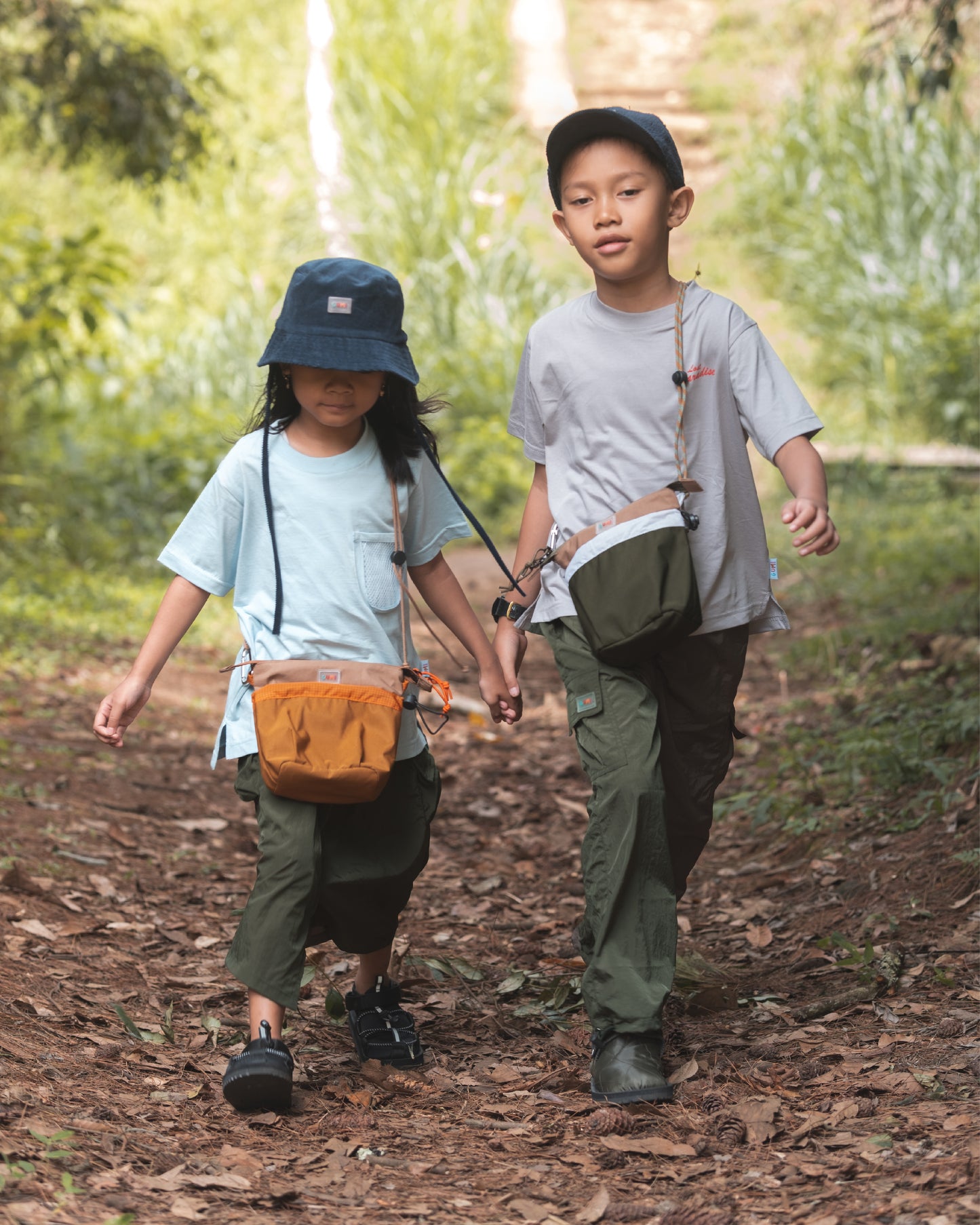 Two children enjoying an outdoor adventure wearing the PIXIES Pumpkin Puddle bag while walking along a nature trail.