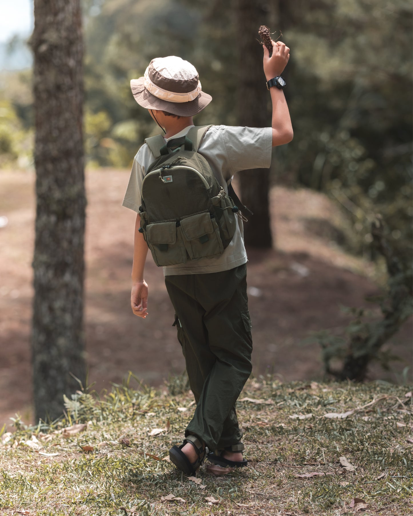 Child exploring nature with the VENEX Dewy Drip Mini Backpack for Kids, wearing a light t-shirt and bucket hat, highlighting the backpack's practical features in a scenic outdoor setting.